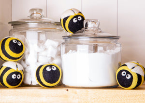 5 bee dryer balls around two glass containers that hold a white laundry powder soap. The bee dryer balls are black and yellow and have fun white googley eyes